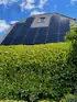 Hausdach mit grauen Ziegeln, Solarmodulen, einem Dachfenster und einem grünen Busch im Vordergrund unter blauem Himmel.